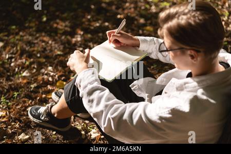 Short hair woman sitting in park on ground and writing notes in her diary. Stock Photo