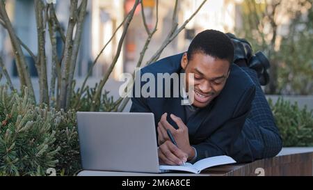Smiling African American student trainee man lying on bench with laptop making notes in notebook outdoors. Funny businessman employee worker working Stock Photo