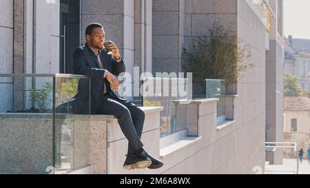 Pensive African American man business worker sitting outdoors near office company city building thinking about work drink tea enjoy work break. Ethnic Stock Photo