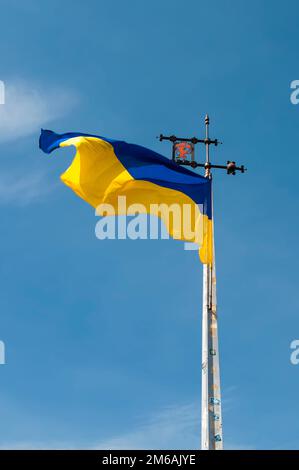 Flag of Ukraine with the coat of arms of Lviv on a background of blue sky, a metal spire with a weather vane. Yellow and blue flag in the wind. Stock Photo