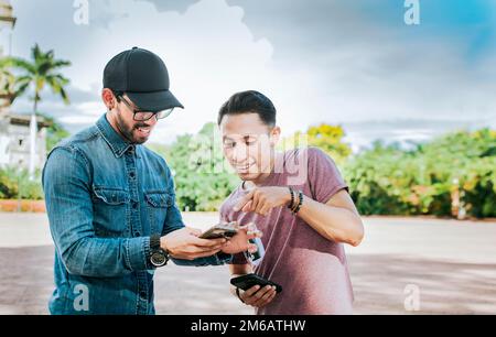 Two young friends having fun with the cell phone outdoors. Two happy friends looking at a cell phone in the street, Two smiling friends looking at Stock Photo