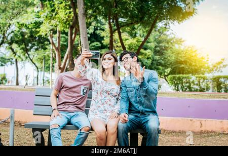 Three friends sitting on a bench taking a selfie, Three teenage friends sitting on a bench taking selfies. Gathering of three happy friends taking a Stock Photo