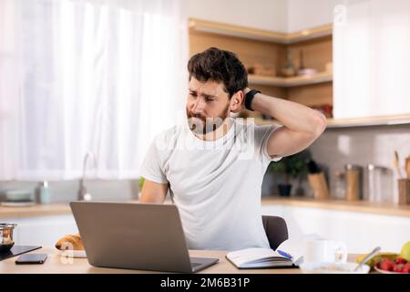 Confused millennial caucasian man with beard scratching his head, works at computer at table in kitchen interior Stock Photo