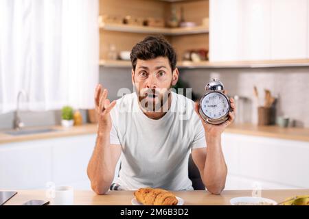 Surprised shocked millennial caucasian guy with beard in domestic clothes with alarm clock late for meeting Stock Photo