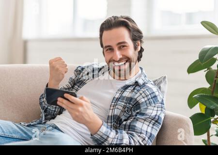 Satisfied young caucasian guy on sofa with smartphone making victory sign with hand, enjoy success in win Stock Photo