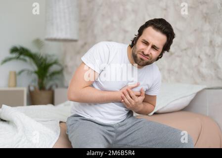 Despaired sad young caucasian guy suffers from chest pain, heart attack, sits on white bed alone in bedroom Stock Photo