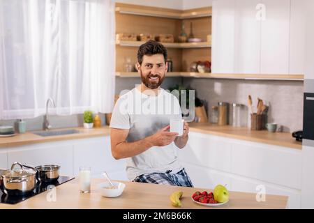 Smiling handsome millennial caucasian guy with beard in domestic clothes with cup of coffee enjoys good morning Stock Photo