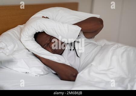Irritated Black Man Lying In Bed And Covering Ears With Pillow Stock Photo
