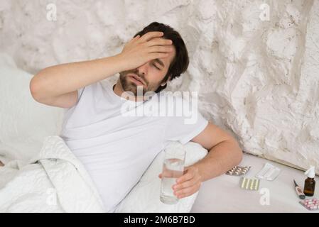 Despaired sad young caucasian man lies on bed with pills, suffers from headache, touch face with hand Stock Photo