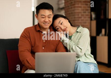 Loving asian couple using laptop together, surfing internet or watching movie online, wife leaning on husband's shoulder Stock Photo