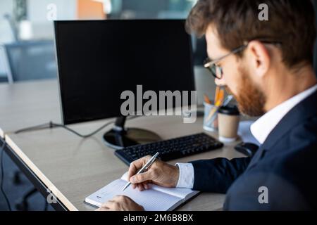 Online business class. Male office worker writing in notebook, taking notes and using computer with black screen, mockup Stock Photo
