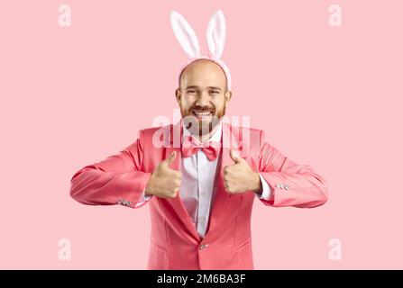 Funny young man in pink suit and Easter bunny ears smiling and showing thumbs up Stock Photo