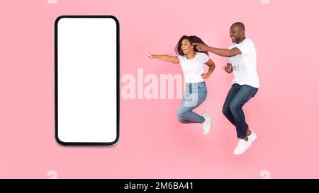 Overjoyed african american spouses jumping in air, pointing fingers at cellphone with blank screen, mockup Stock Photo