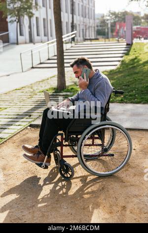 Man in wheelchair talking on the phone while working with laptop outdoors. Stock Photo