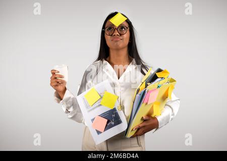 Busy tired young black business woman in glasses with many folders with cup coffee takeaway look at sticker on forehead Stock Photo