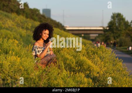 Beautiful African girl with curly hair drinking beer in a field of yellow flowers Stock Photo