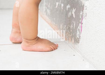 close-up of a brunette baby's feet, standing on a white tile, next to a wall with damaged paint. Stock Photo