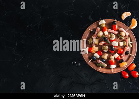Snack skewers on a wooden tray on a black concrete table. Appetizers with mozzarella, ham, olives, cucumbers, tomatoes. Stock Photo