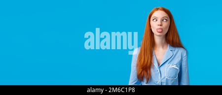 Be funny and silly. Redhead carefree and enthusiastic caucasian girl squinting, making goofy expression, showing tongue, standing blue background in Stock Photo