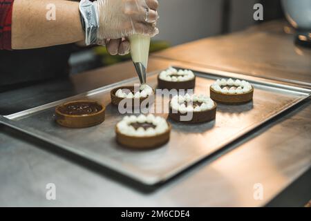 A baker wearing transparent glove decorating tarts placed on a baking tray with white cream using a pastry bag. Blurred background. High quality photo Stock Photo