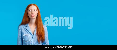 Be funny and silly. Redhead carefree and enthusiastic caucasian girl squinting, making goofy expression, showing tongue, standing blue background in Stock Photo