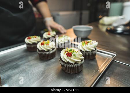 Side view of six cupcakes decorated with white cream and colorful sparkles placed on a baking tray. A person wearing a transparent glove and black apron standing in the kitchen blurred in the background. High quality photo Stock Photo