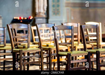 A close up view of wooden chairs with green wicker seats lined up in front of a rack holding lit votive candles. Stock Photo