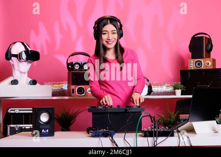 Asian musician mixes electronic techno sounds, wearing headphones at DJ table in club. Performing live, using turntables for an unforgettable concert experience. Surrounded by fans at the party Stock Photo