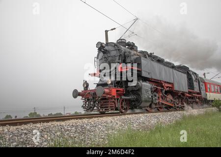 Vintage steam engine locomotive speeding on railroad tracks curve and blowing heavy white smoke. Stock Photo