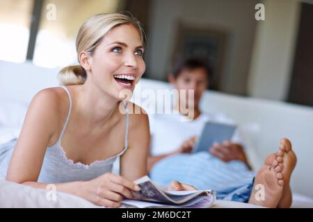 Moments of marital bliss. a happy young woman lying in bed and reading a magazine while her husband uses a digital tablet in the background. Stock Photo