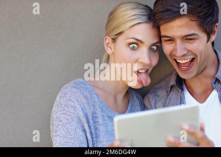 Getting their silly on. a playful young couple making funny faces together while using a digital tablet against a gray background. Stock Photo