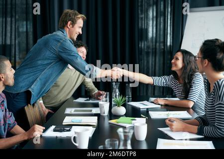 They always make new team members feel welcom. two colleagues shaking hands in a boardroom while coworkers look on. Stock Photo