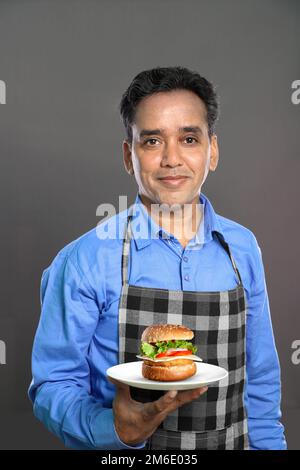 Indian Male Chef with a Burger in Hand on Grey Background Stock Photo
