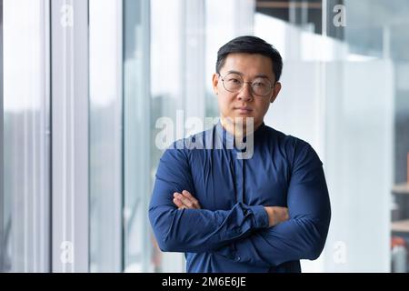 Portrait of asian businessman inside modern office, boss in glasses serious looking at camera, man with crossed arms thinking worker. Stock Photo