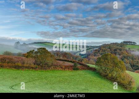 An early morning mist rising from a beautiful patchwork of undulating farmland in autumn. The blue sky is broken by small floating white clouds. Stock Photo