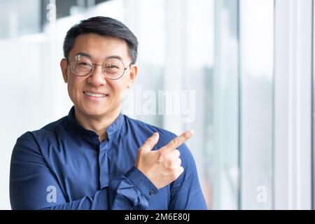 Portrait of happy and successful asian businessman, man smiling and looking at camera pointing with finger towards window, satisfied with achievement worker. Stock Photo
