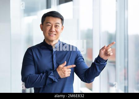 Portrait of happy and successful asian businessman, man smiling and looking at camera pointing with finger towards window, satisfied with achievement worker. Stock Photo