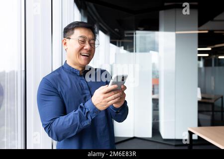 Satisfied with achievement Asian businessman inside office using phone, mature man in shirt near window reading news online from smartphone, successful boss in glasses. Stock Photo