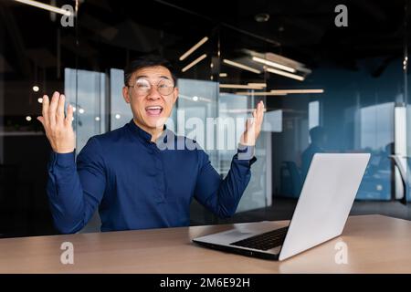 Successful asian businessman celebrating victory success, portrait of man in office in casual shirt, employee smiling and looking at camera satisfied with achievement holding hand up victory gesture. Stock Photo