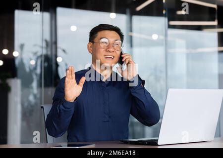Successful and cheerful asian businessman talking on the phone with clients inside the office, mature man in a shirt working on a laptop at the workplace. Stock Photo