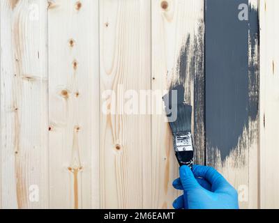 Hand of a worker in blue rubber gloves paints a wooden wall with a brush of gray paint Stock Photo