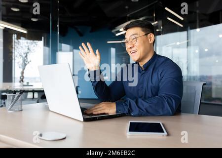 Video call happy asian talking remotely with colleagues, businessman inside office with laptop on online meeting with employees, sitting at desk at workplace man in shirt. Stock Photo