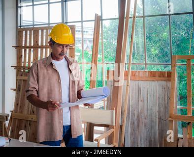Portrait of a senior carpenter looking at blueprints plans, rechecking and comparing drawing plans. Hobby, Retirement Concept Stock Photo