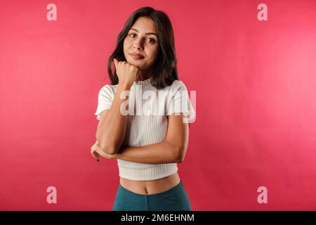 Brown-haired woman wearing white ribbed crop isolated over red background touches chin with fist and looks very bored. Doesn't believe the lies and wa Stock Photo
