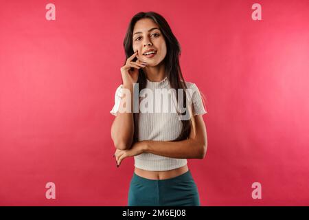 Cute brunette woman wearing white ribbed crop isolated over red background enjoys talking and laughs a lot. He touches his chin with his hand and like Stock Photo