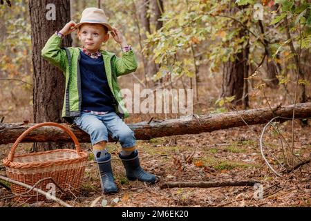 Little boy in a hat in the autumn forest. Boy with a basket in the woods. Little forester. Near a fallen tree. Stock Photo