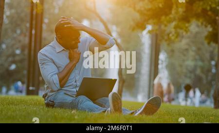 Elderly middle-aged African American businessman entrepreneur man sitting on grass in city park working on computer laptop feeling tired stressed Stock Photo