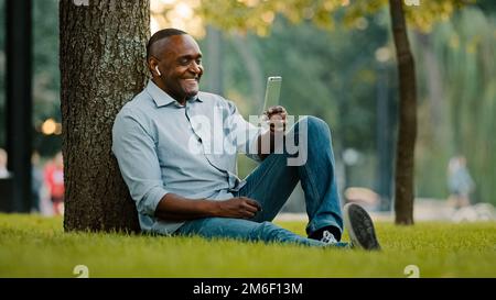 Elderly ethnic man African American smiling friendly entrepreneur sitting on grass in city park businessman with smartphone talk video call chat Stock Photo