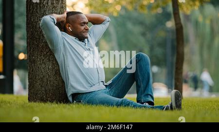 Calm relaxed resting middle-aged male listening music with earphones sitting on green grass in park under tree enjoy song relaxing adult African Stock Photo