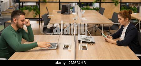 Red-haired caucasian woman uses a smartphone and a bearded man works behind a laptop in an open space office.  Stock Photo